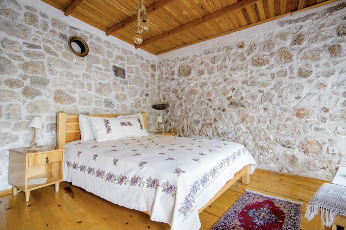 Un dormitorio con pared de piedra, techo de madera, cama con colcha estampada de flores, mesita de noche y alfombra roja con diseño.