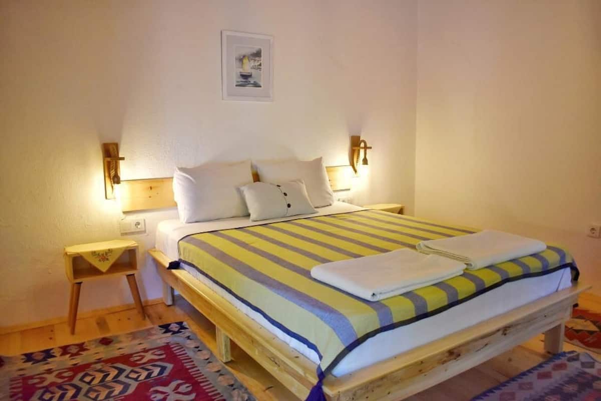 Una cama individual con una colcha a rayas y dos almohadas, una mesita de noche con una lámpara, y una pequeña alfombra en el suelo.