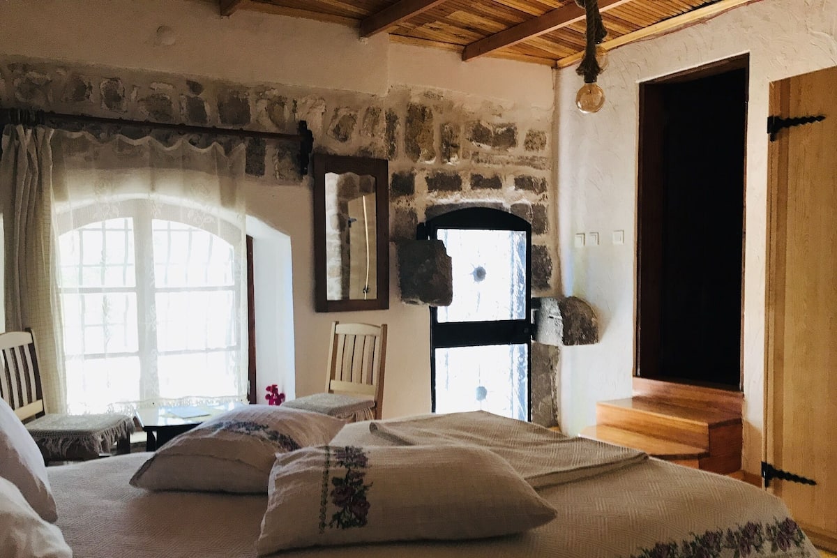 Una habitación rústica con pared de piedra, techo de madera, una cama con almohadillas florales, una silla y una ventana con vistas.