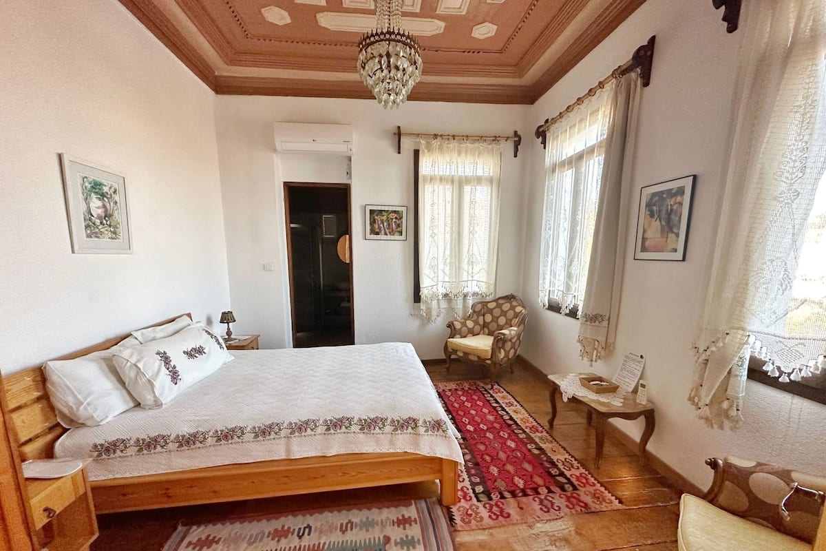 Un dormitorio con una cama de madera, una silla, una mesita, una araña y una puerta.