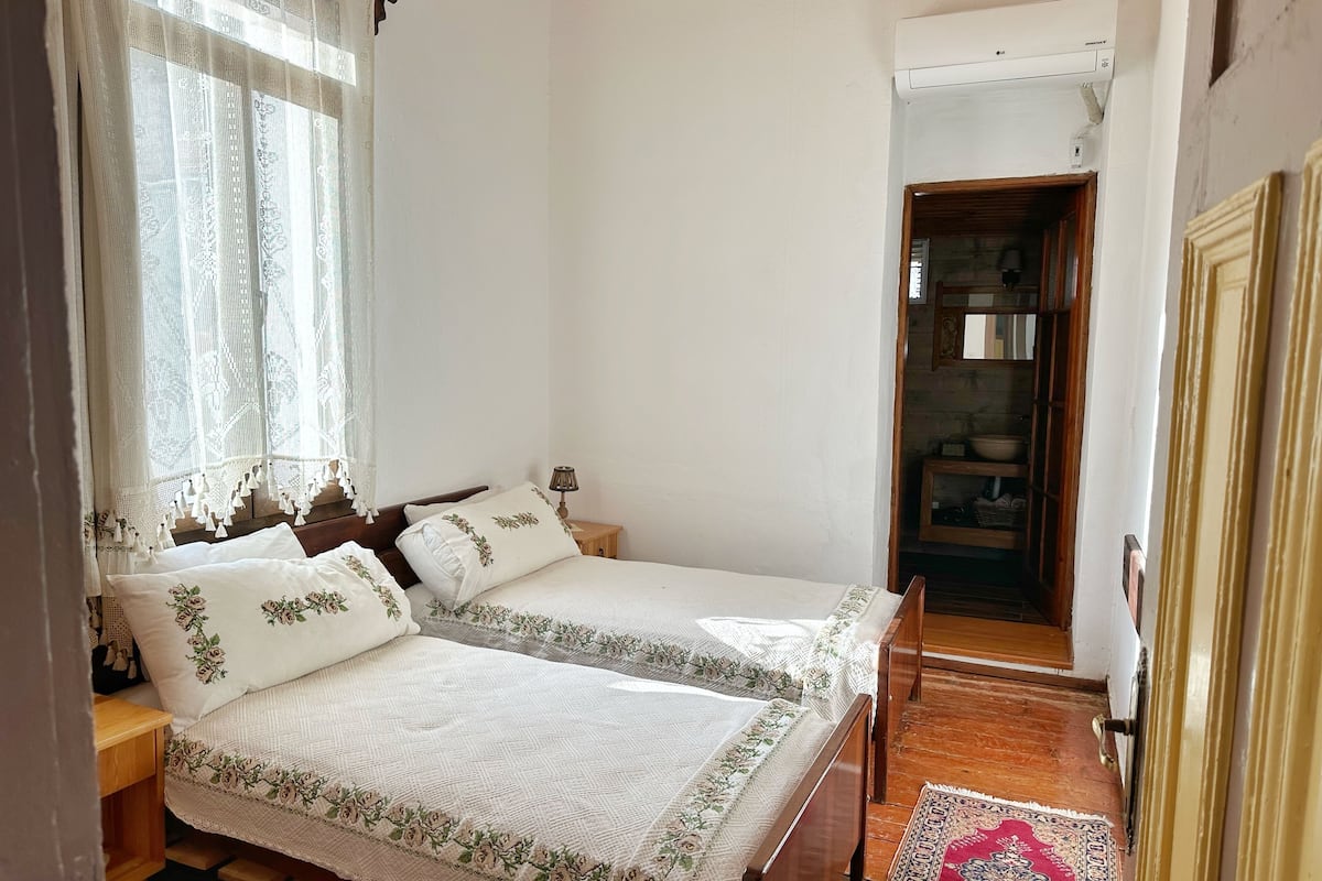 Un dormitorio con dos camas individuales, una mesita de noche de madera, una lámpara de noche, una ventana con cortinas de encaje y una puerta de madera que da a un baño.