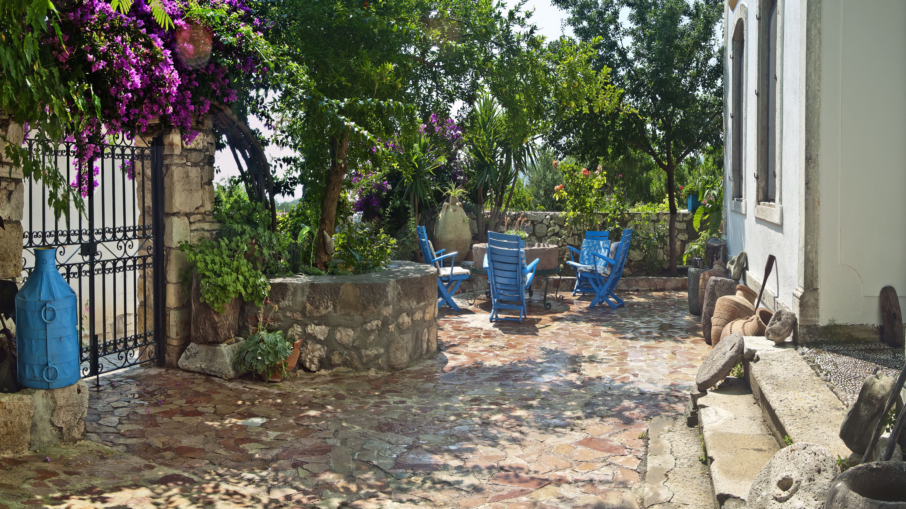 Un patio empedrado con sillas azules y una mesa, rodeado de abundante vegetación y plantas en flor.