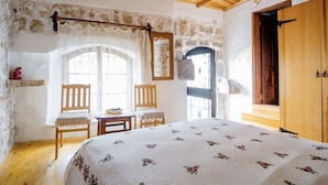 A bedroom with a wooden ceiling, stone walls, a bed with a floral bedspread, two wooden chairs, a small table, and a wooden door. - Saklı Konak (Karaburun)