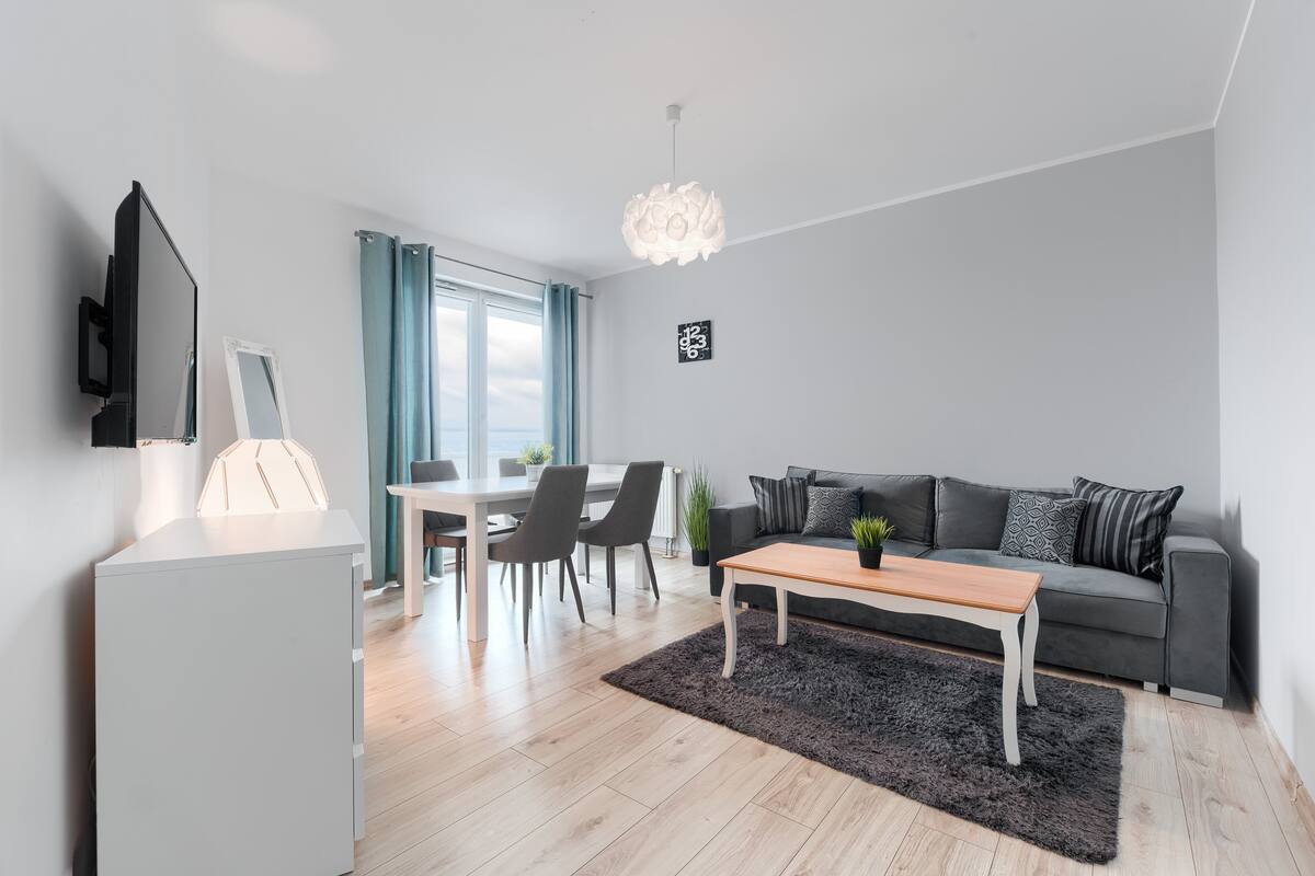 Un salon moderne avec un canapé gris, une table basse en bois, un téléviseur fixé au mur et un coin repas comprenant une table et des chaises.