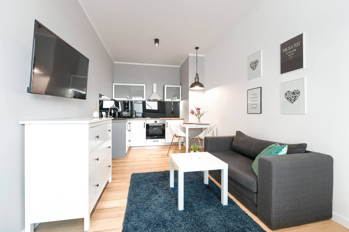 Un salon moderne avec un canapé gris, une table basse blanche et un tapis bleu. On aperçoit, au fond, une cuisine avec des meubles blancs et un téléviseur.