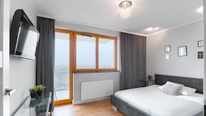 Een moderne slaapkamer met een groot bed, een tv aan de muur, een glazen bijzettafeltje met planten, uitzicht door een groot raam en een radiator.