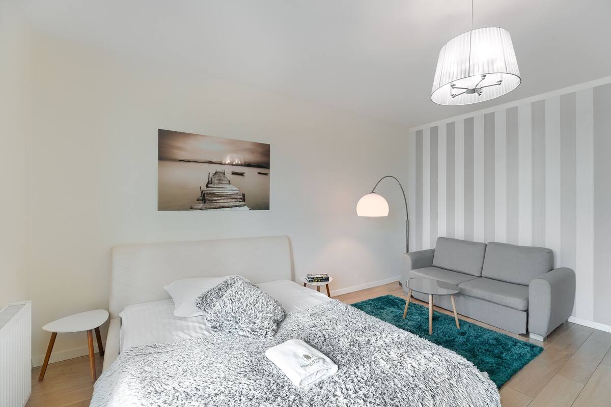Une chambre à coucher moderne avec un lit, un canapé gris, une petite table, une lampe et un tableau au mur.