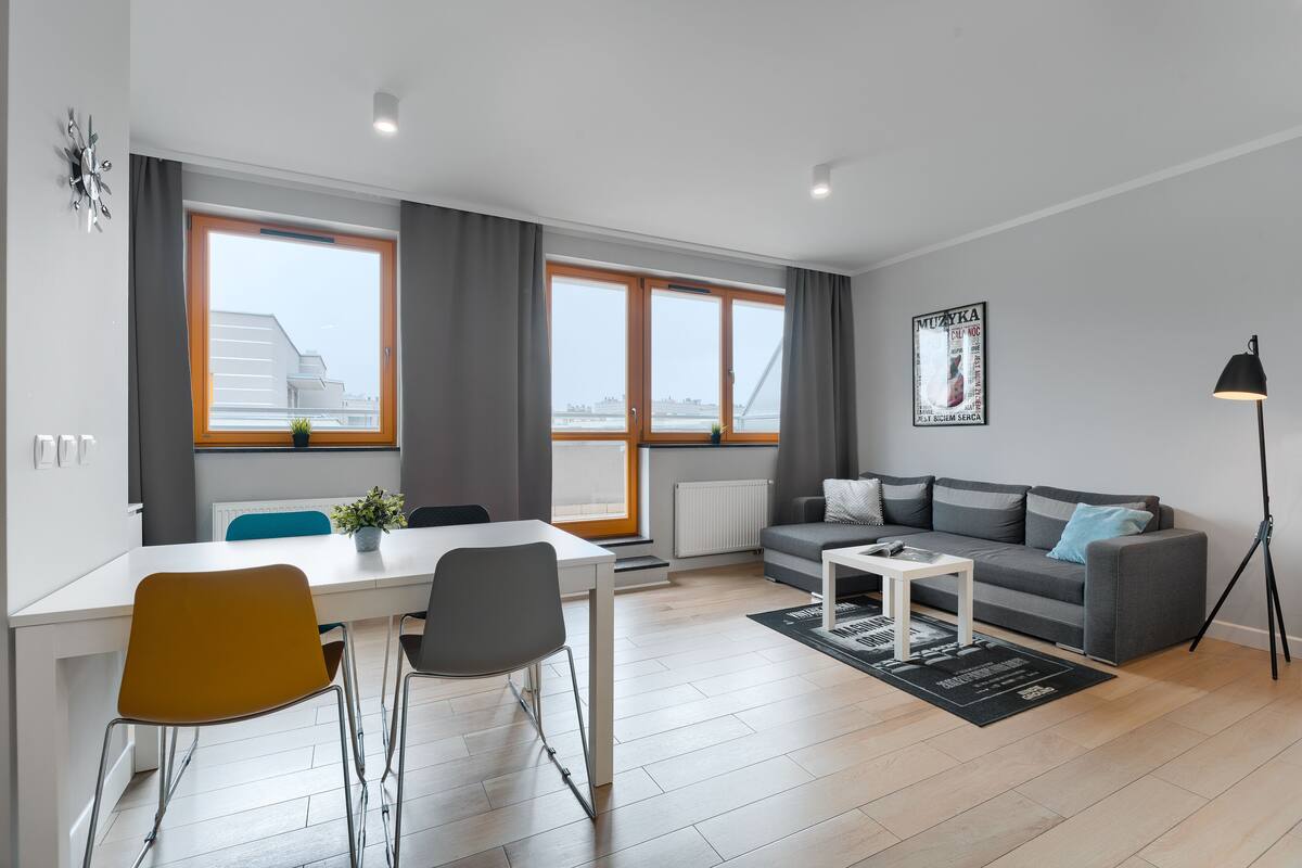 Un salon moderne avec un canapé gris, une table à manger avec deux chaises, un lampadaire et de grandes fenêtres avec des rideaux.