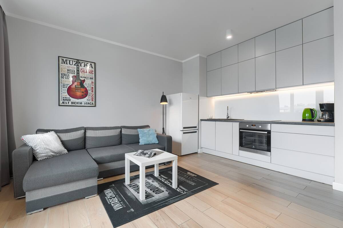 Un salon moderne avec un canapé gris, une petite table blanche et un coin cuisine doté de meubles blancs et d’appareils électroménagers blancs.