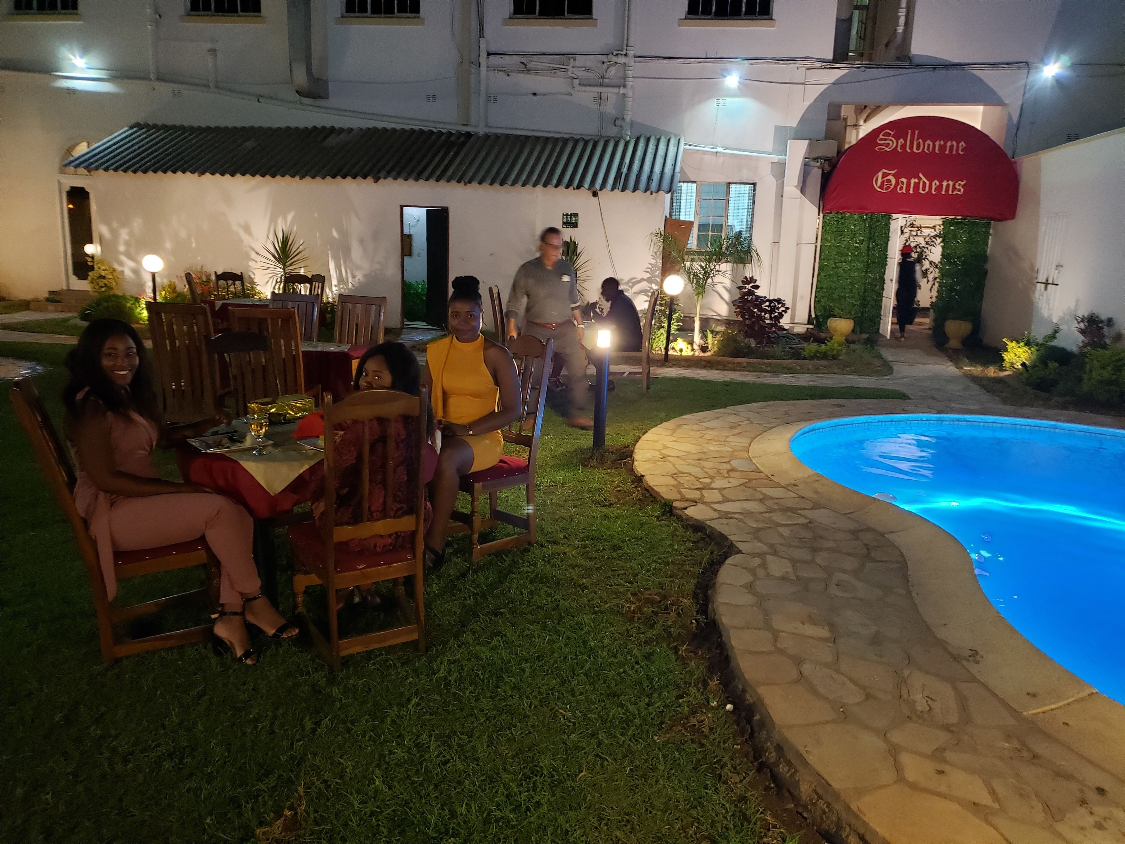 Un área de comedor al aire libre con mesas y sillas, una piscina y un edificio con un letrero que dice "Selborne Gardens".