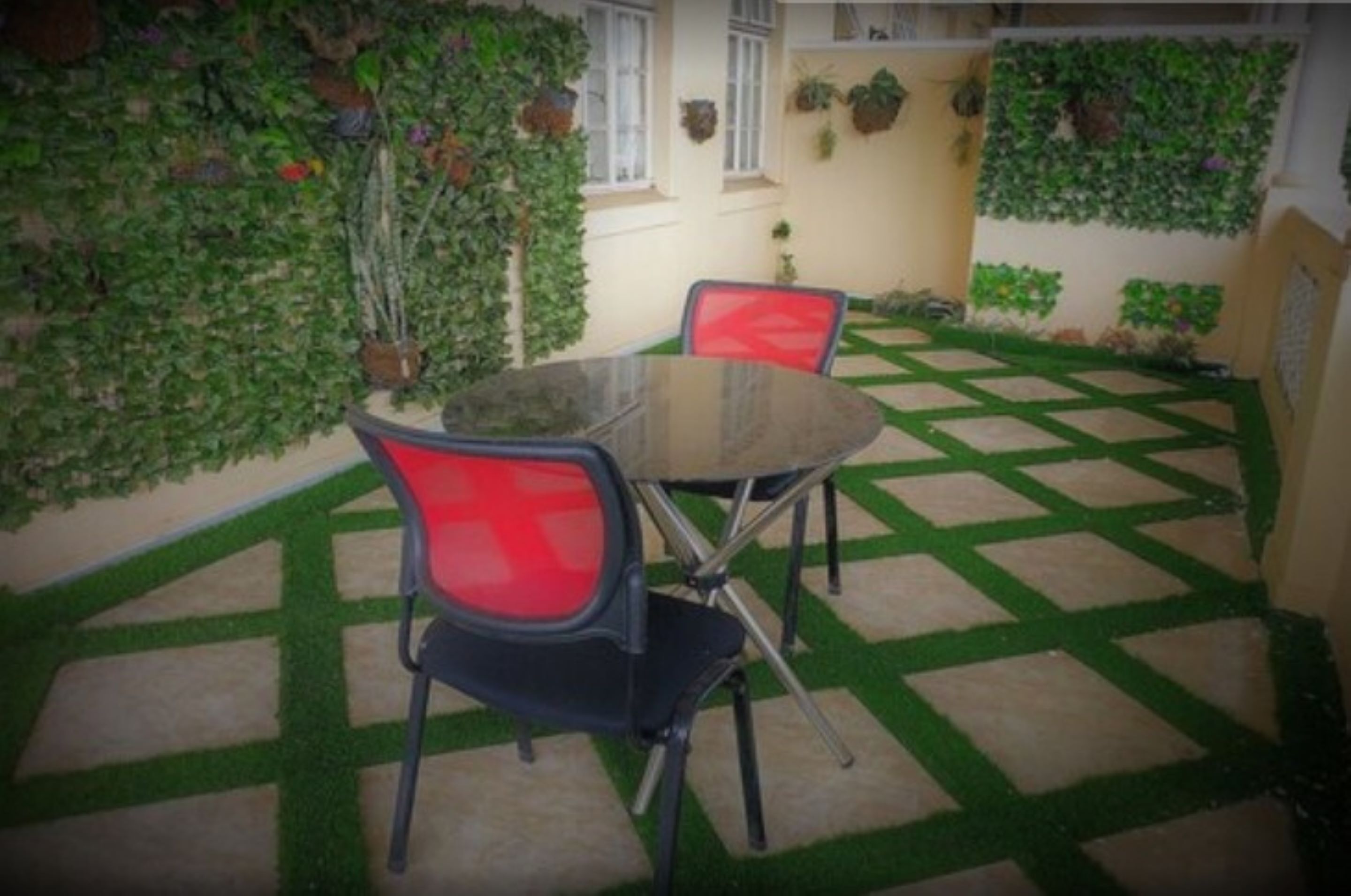 A small outdoor patio with a glass table and two red chairs, surrounded by a green and beige tiled floor and walls covered with climbing plants.