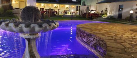 A fountain with water cascading into a pool, surrounded by stone tiles and a well-lit outdoor seating area.