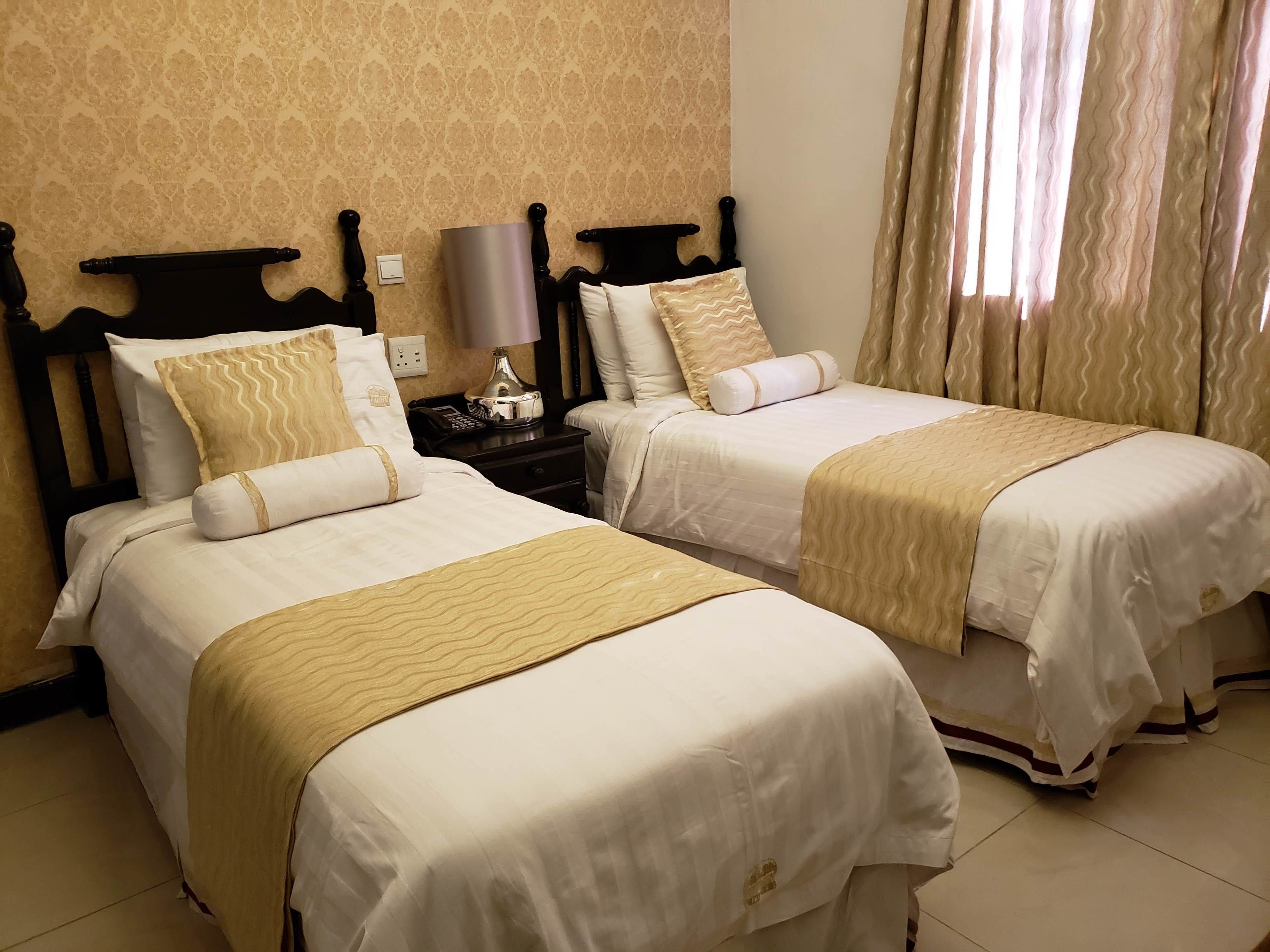A hotel room with two single beds, a nightstand with a lamp, a telephone, and a wall socket.