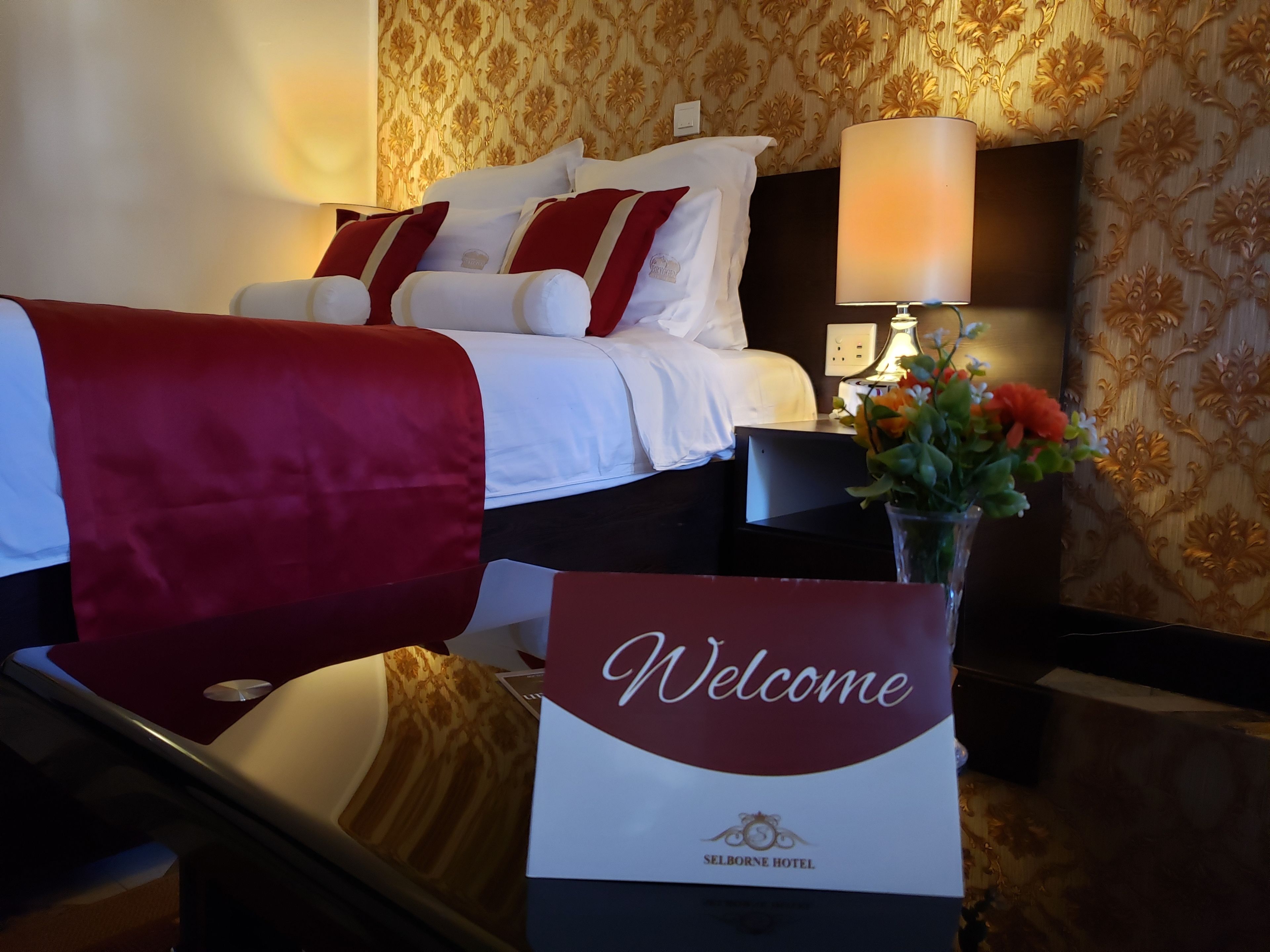 A hotel room with a bed, a nightstand with a lamp, and a welcome sign.