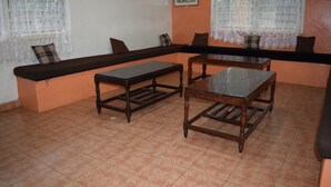 A living room with a sofa set, two wooden tables, and lace curtains on windows. - Mountainview Backpackers Kisumu (Kisumu)