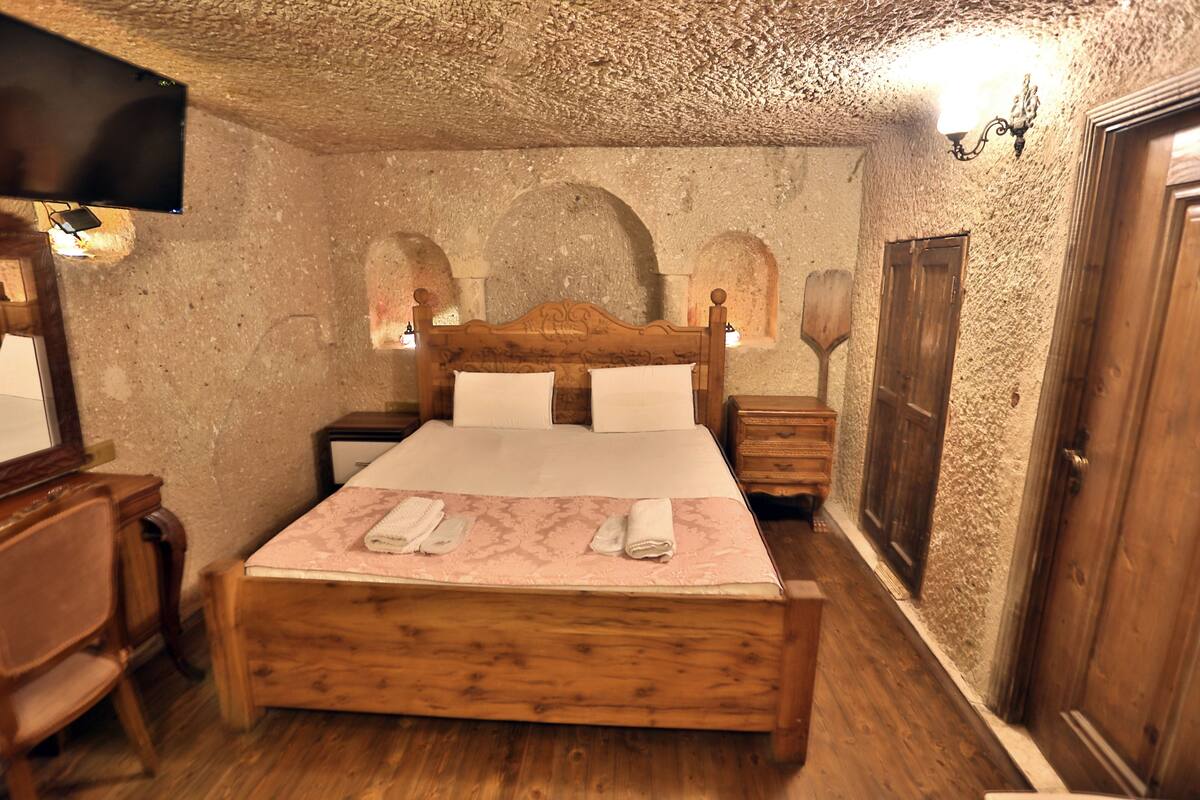 A bedroom with a wooden bed, a wooden nightstand, a wooden chair, a flat-screen TV, and a wall-mounted light fixture.