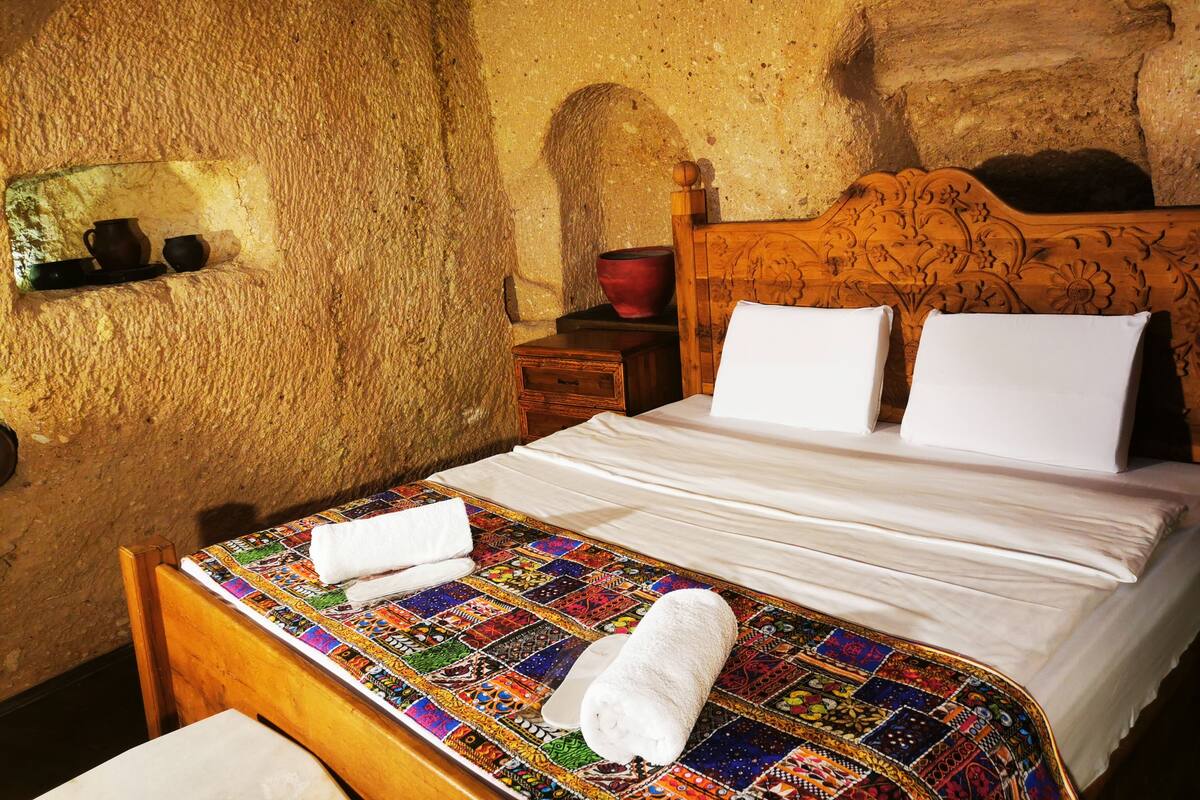 A room with a stone wall, a wooden bed with a patterned bedspread, a wooden nightstand, and a red vase.