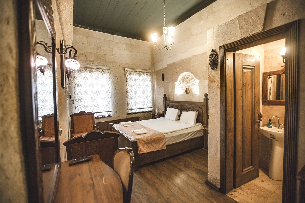 A room with a wooden bed, a desk, and a chair. There is a window with lace curtains and a chandelier.