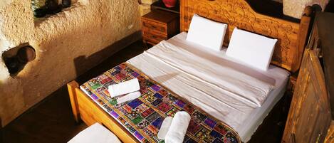 A rustic stone-walled room with a wooden bed, a small wooden nightstand, and a colorful woven mat on the floor.
