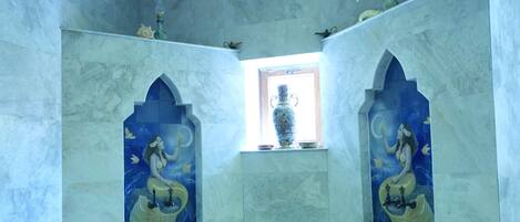 A room with a high ceiling, a skylight, and a central window. There are two wall niches with decorative figures, a small window with a vase, and a stone bench with bowls.