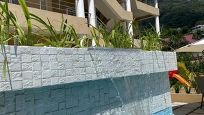 A swimming pool with a stone wall and a view of a building with balconies and a mountain in the background. - Tropical Hideaway (Mahé Island)