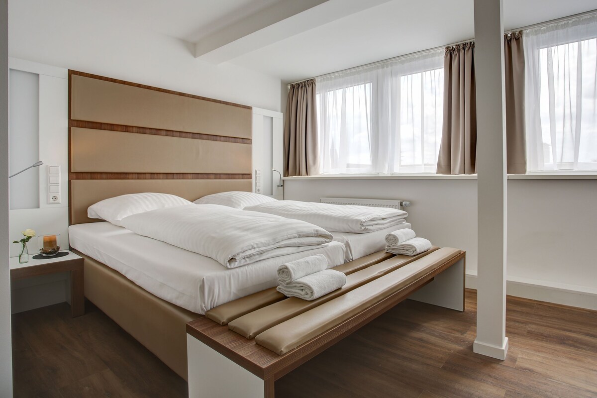 Una habitación de hotel moderna con una cama grande, una mesita de noche con lámpara y una ventana con cortinas.