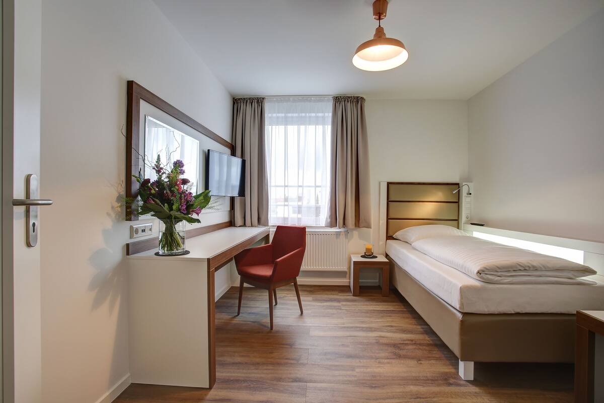 A bedroom with a bed, a desk, a chair, a TV, a window with curtains, and a small bedside table with a lamp.