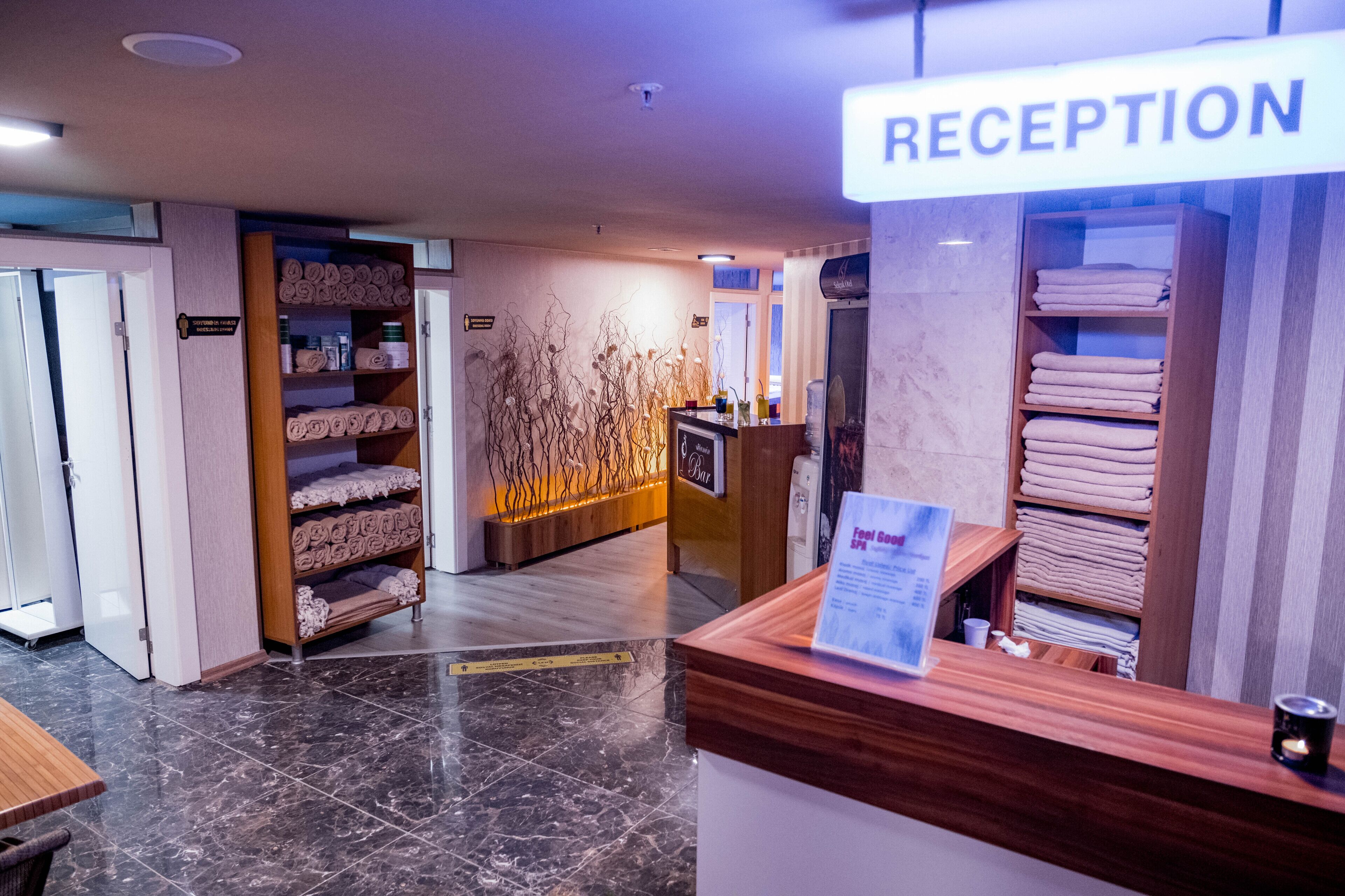 A reception area with a sign labeled "RECEPTION," a wooden reception desk, a counter with a menu, and shelves with towels.