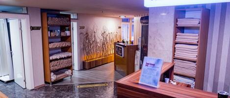 Zona de recepción con un cartel que dice "RECEPTION", un mostrador de madera, un mostrador con un menú y estantes con toallas.