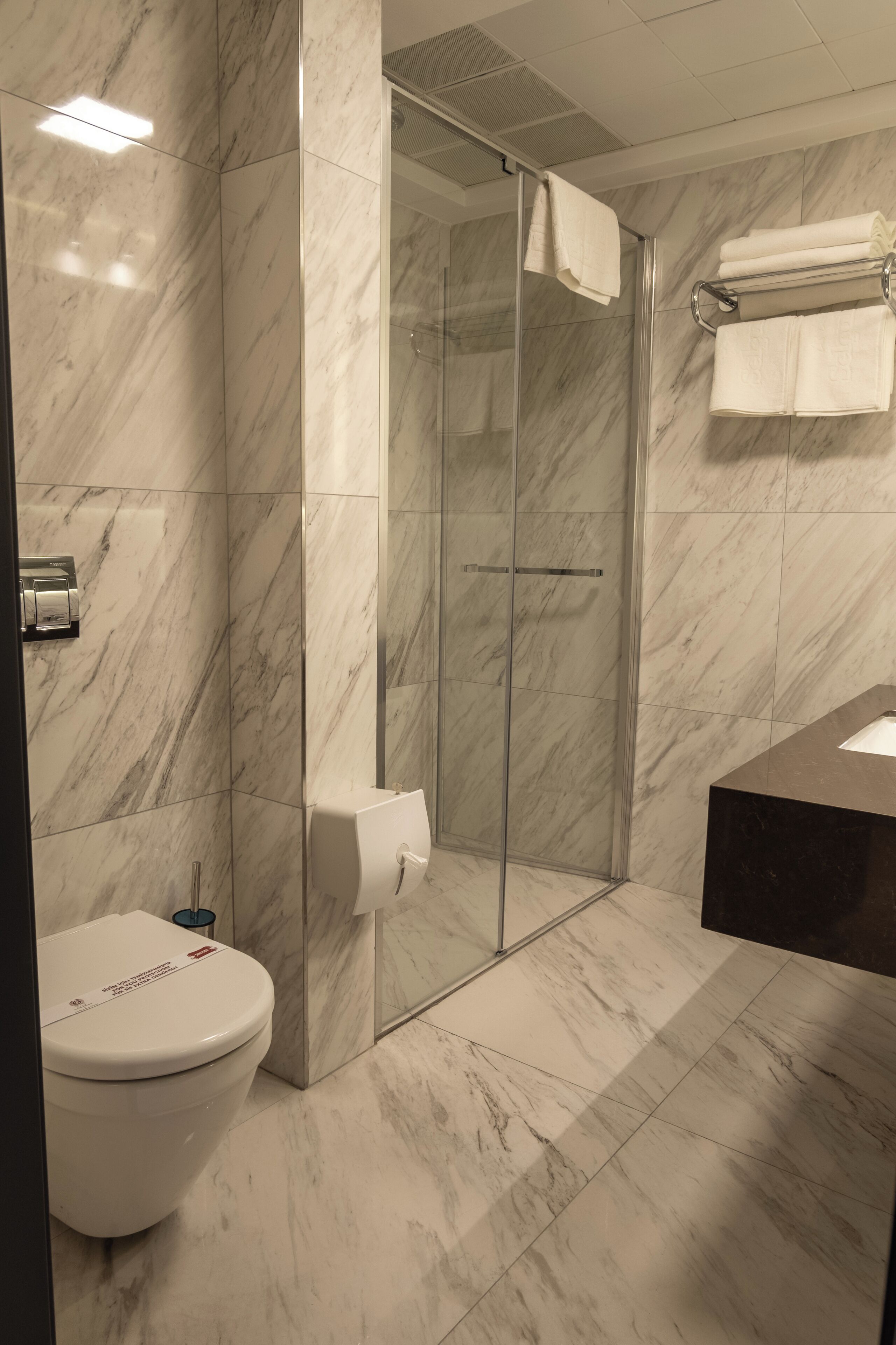 Een badkamer met een marmeren vloer, toilet, een glazen douchecabine en een handdoekrek met handdoeken.