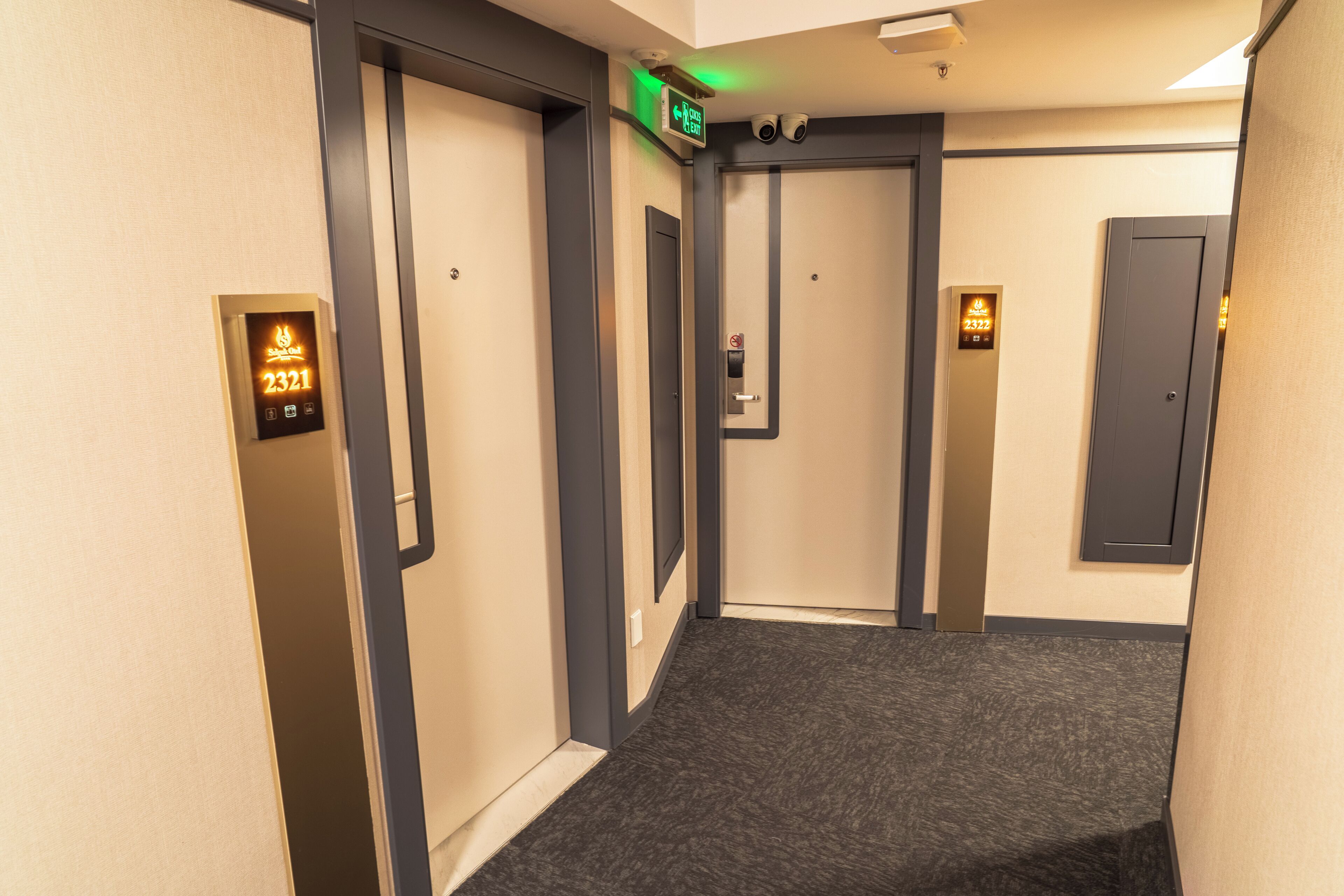 Un couloir d’hôtel avec un sol recouvert de moquette, un écran numérique indiquant l’étage de l’ascenseur et une porte équipée d’une caméra de surveillance.