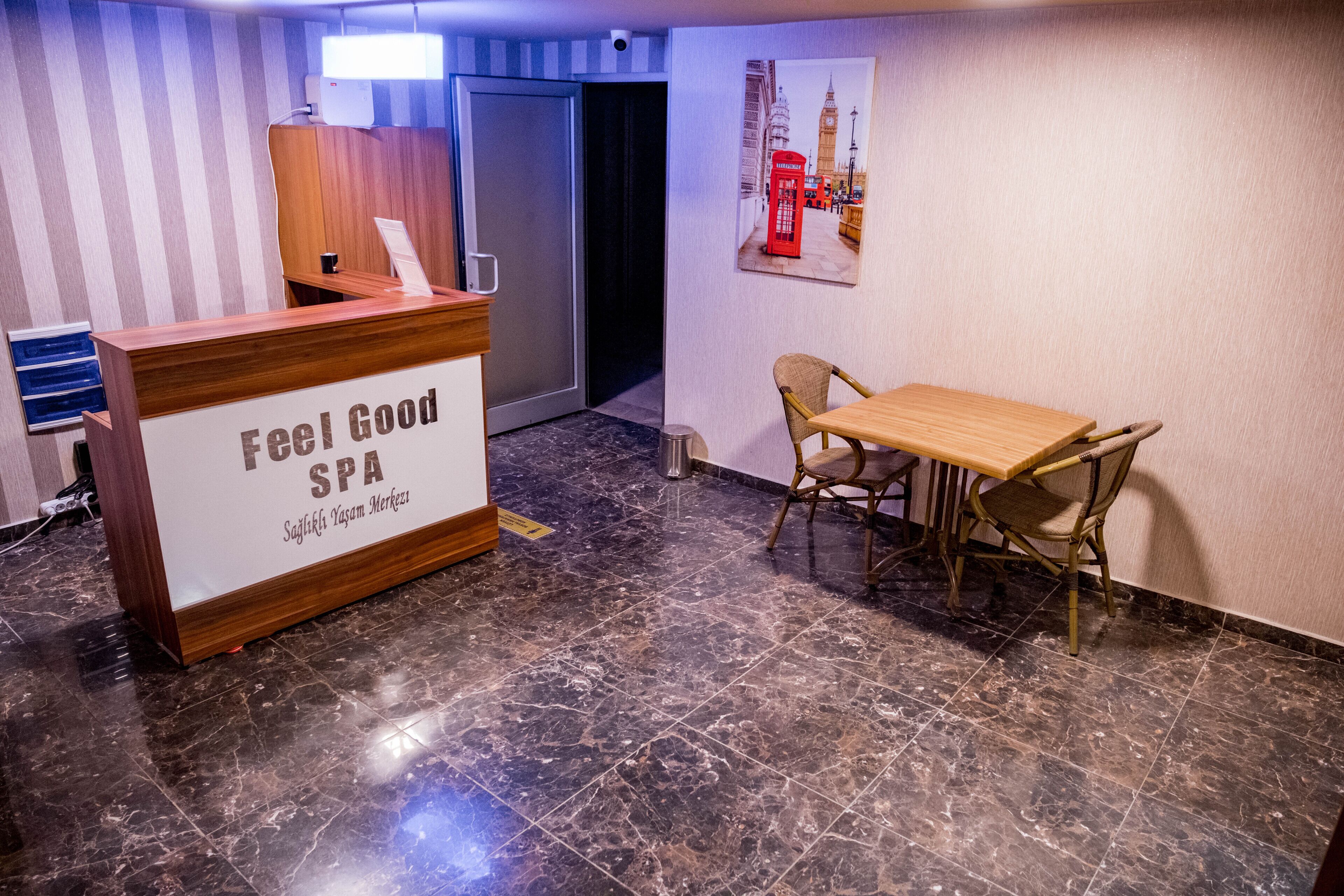 Uma receção com o nome "Feel Good Spa", acabamento em madeira, uma cadeira, uma mesa e um cartaz de uma cabine telefónica vermelha na parede.