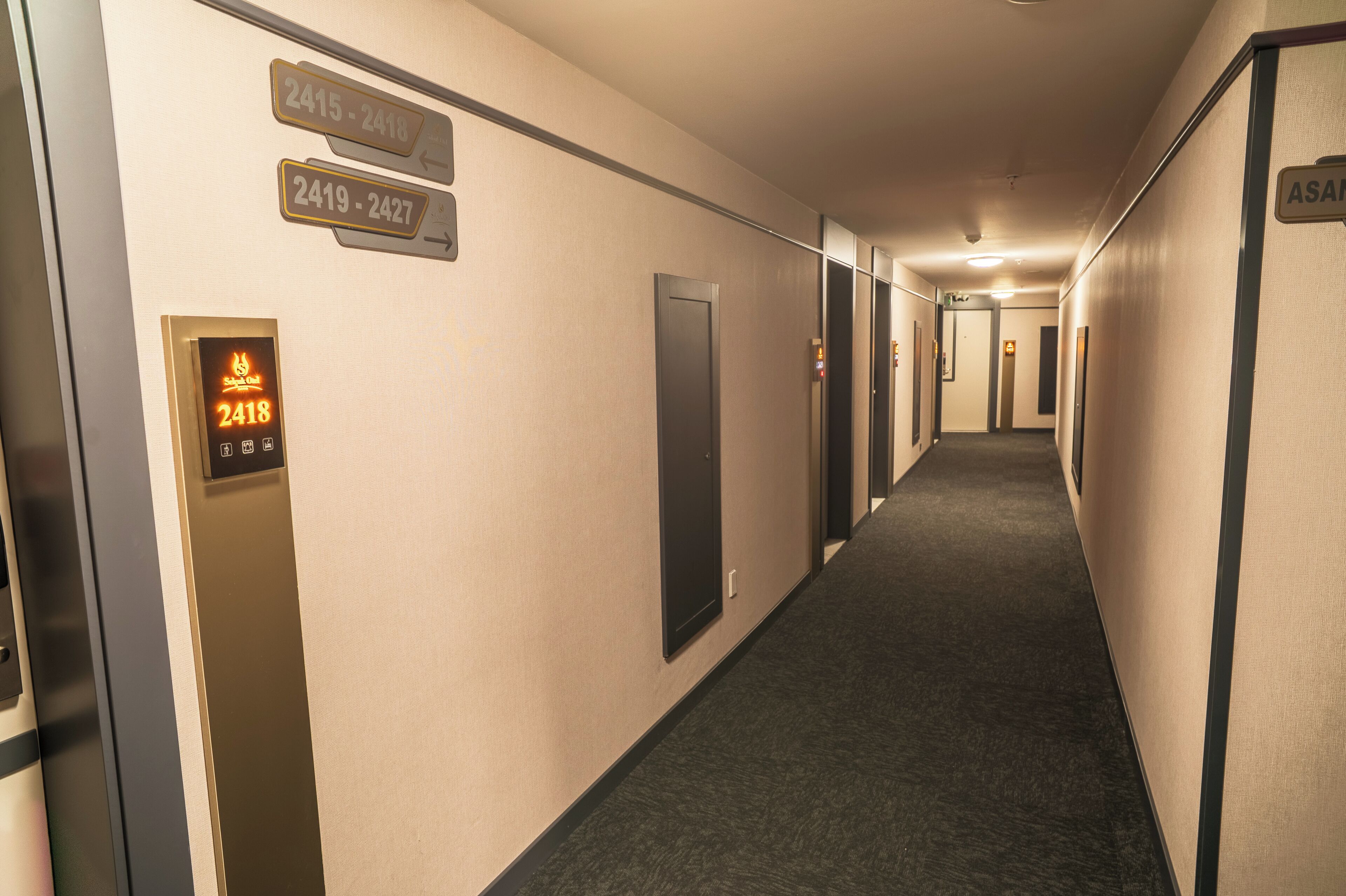Pasillo de hotel con señales de indicaciones, un ascensor y una puerta con el rótulo 'ASAM'.