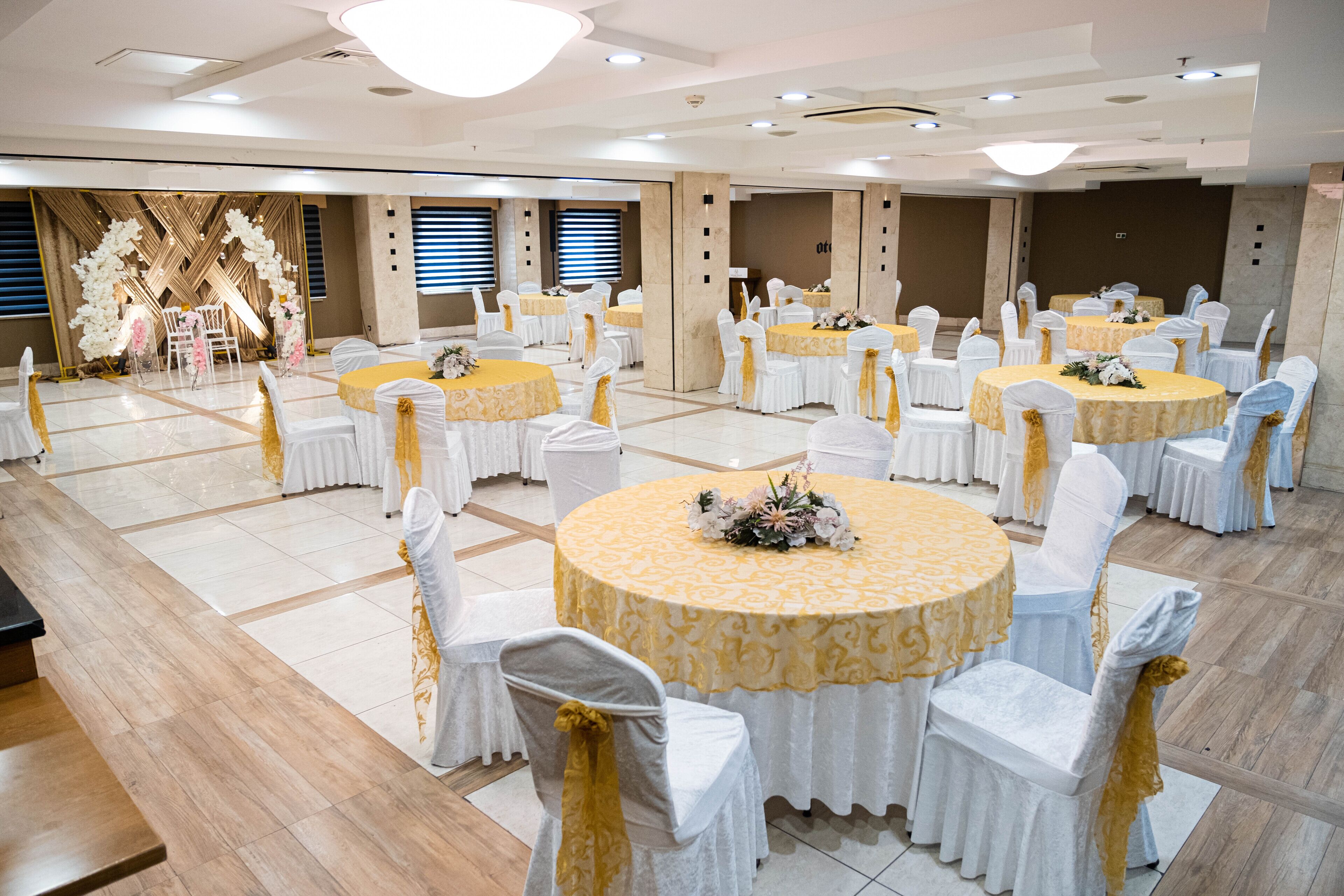 Un salón de banquetes con mesas redondas cubiertas con manteles amarillos, sillas blancas adornadas con cintas doradas y un escenario decorado con flores.