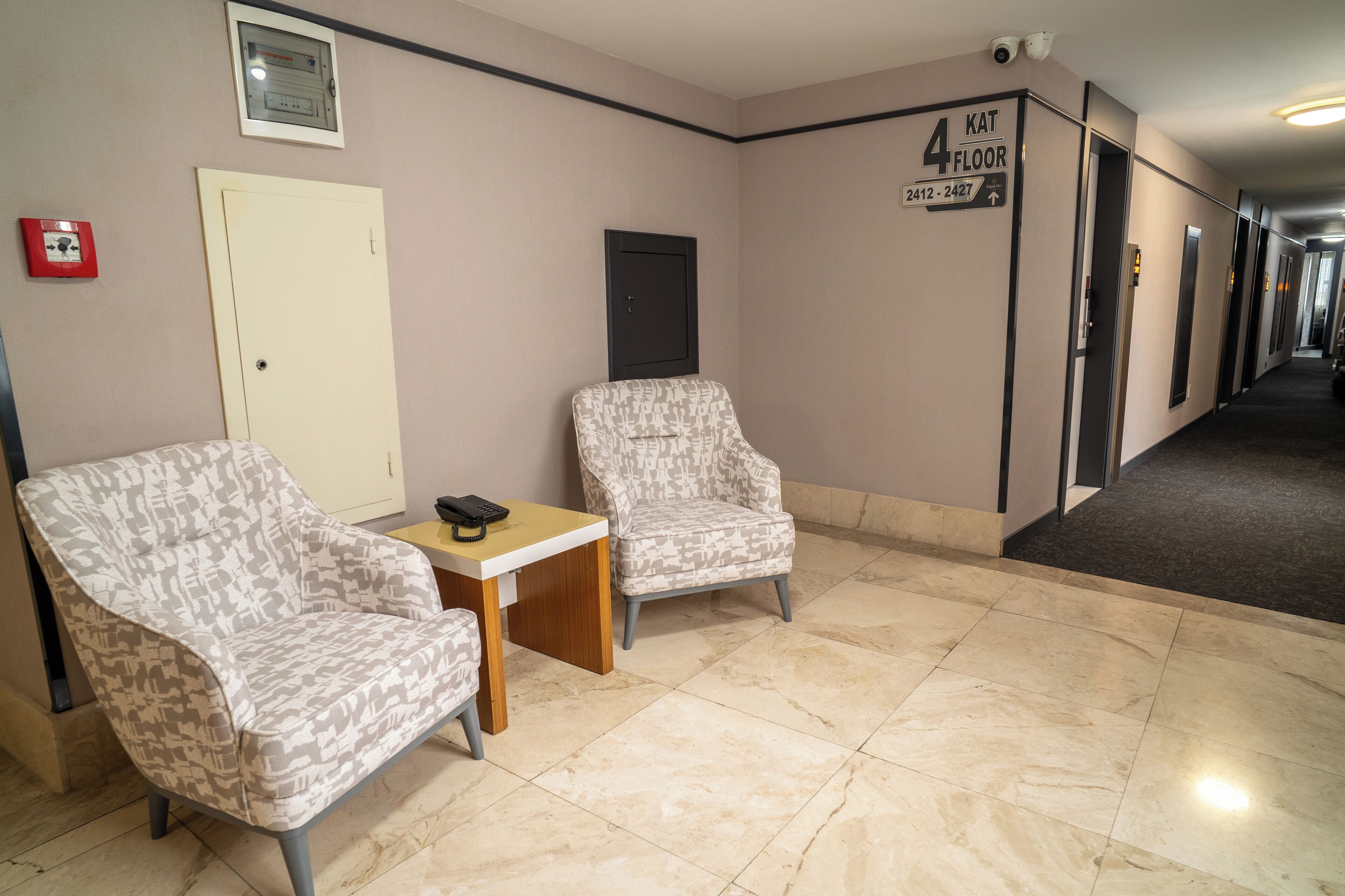 A hotel lobby with two patterned armchairs, a small wooden table, a fire alarm, and a sign indicating the 4th floor.