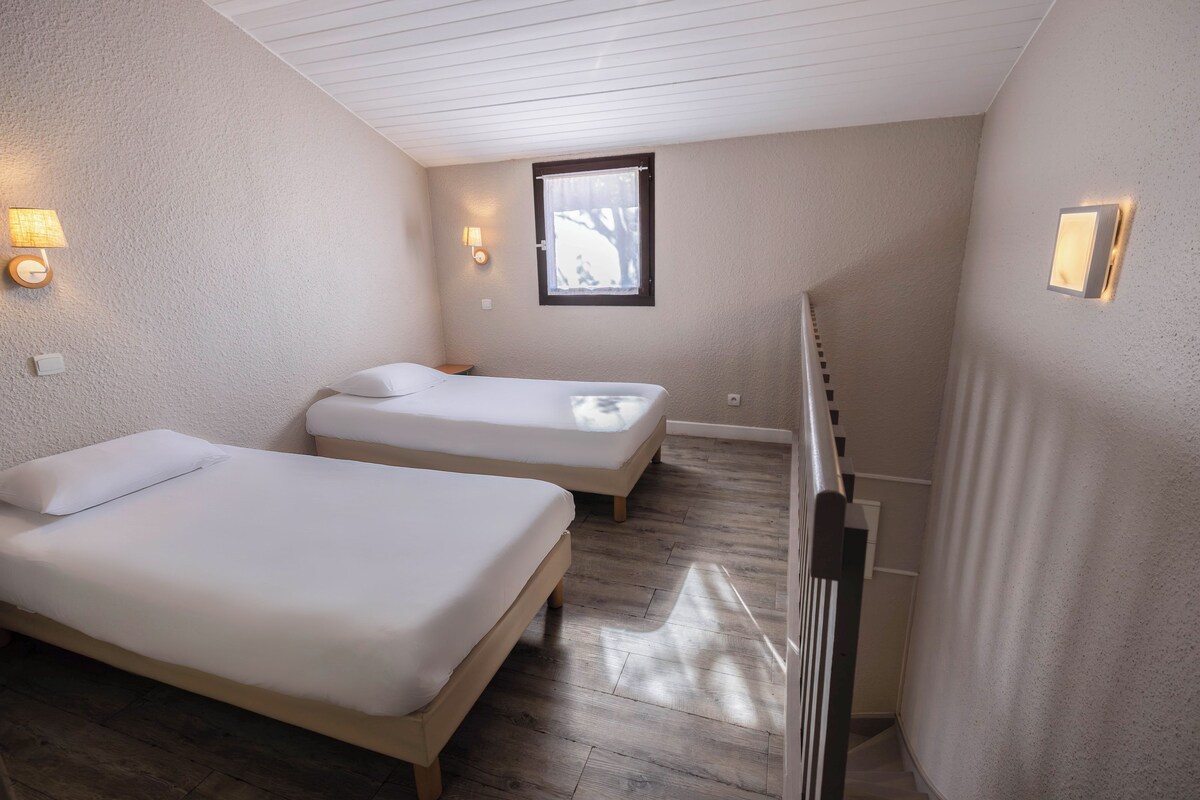 Een ruime, goed verlichte kamer met twee enkele bedden, houten vloeren en een raam met uitzicht op bomen.