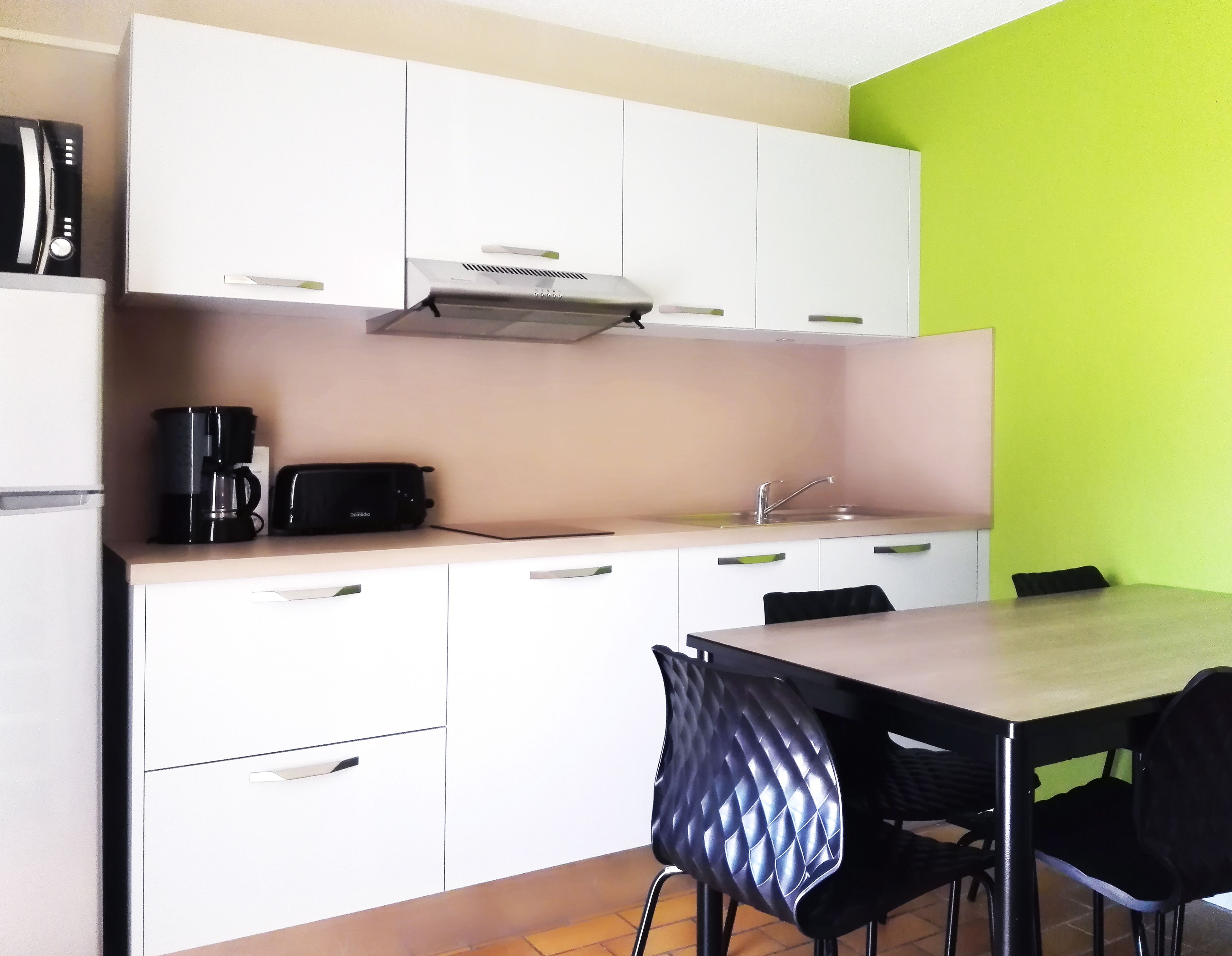 A compact kitchen with white cabinets, a black coffee maker, a toaster, and a sink. There is a dining table with black chairs.