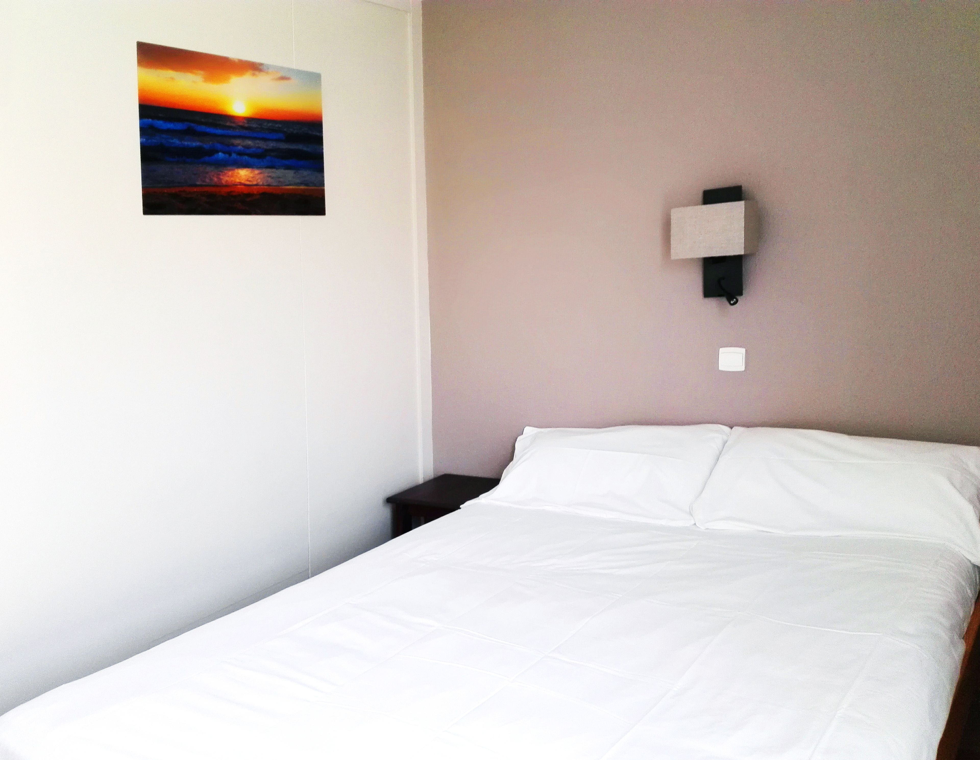 Une chambre d’hôtel avec un lit, une table de chevet, une lampe fixée au mur et un tableau représentant un coucher de soleil.