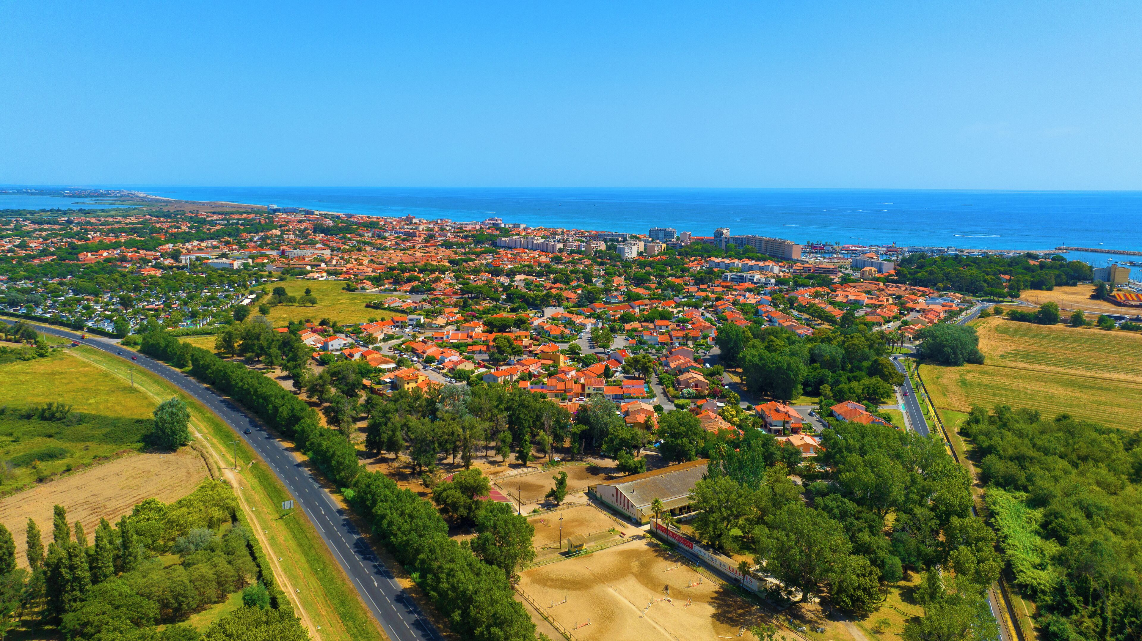 An aerial view of a coastal town with red-roofed houses, a main road, and a beach in the background.