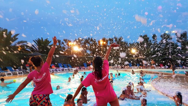 Piscina com pessoas aproveitando a água, cercada por palmeiras e espreguiçadeiras.
