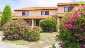 Edifício residencial com telhado de telhas, várias janelas e vegetação exuberante, incluindo arbustos floridos.