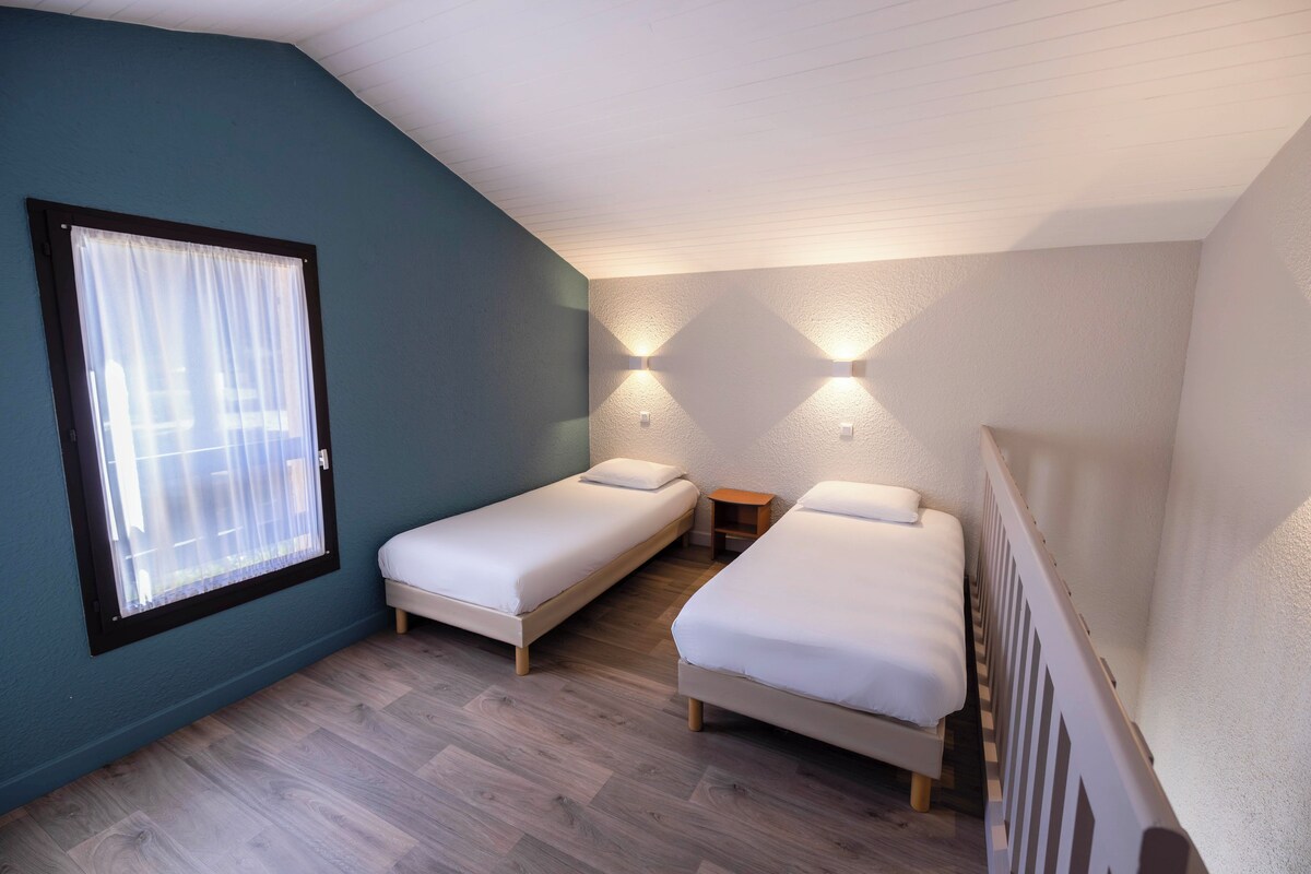 Een kleine, goed verlichte kamer met twee eenpersoonsbedden, een raam met gordijnen en wandlampen.