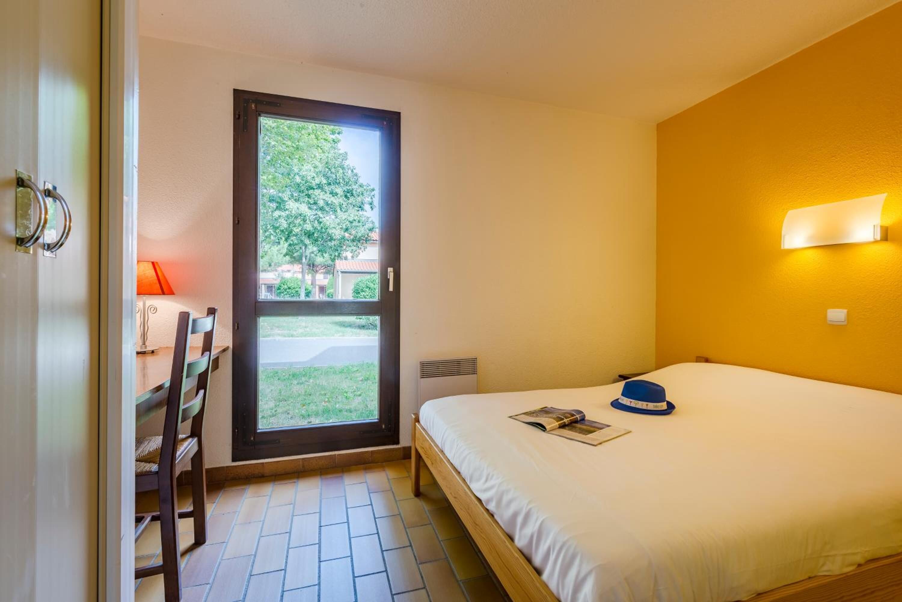 A bedroom with a bed, a desk with a chair, a window, and a wall-mounted light fixture.