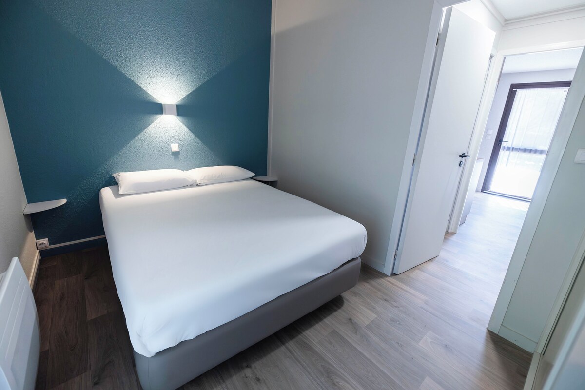 A bedroom with a bed, a nightstand, a wall-mounted light, and a door leading to another room.