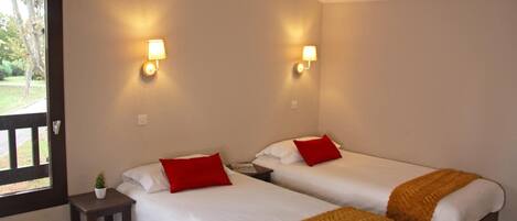 A room with two beds, each with white bedding and red pillows, a small table with a plant, and wall-mounted lamps.