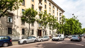 Um edifício de vários andares com fachada bege, carros estacionados na rua e árvores ao longo da calçada.