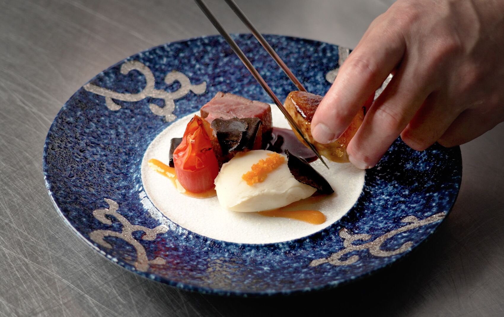 A hand using chopsticks to pick up a piece of food from a blue ceramic plate with a decorative pattern.