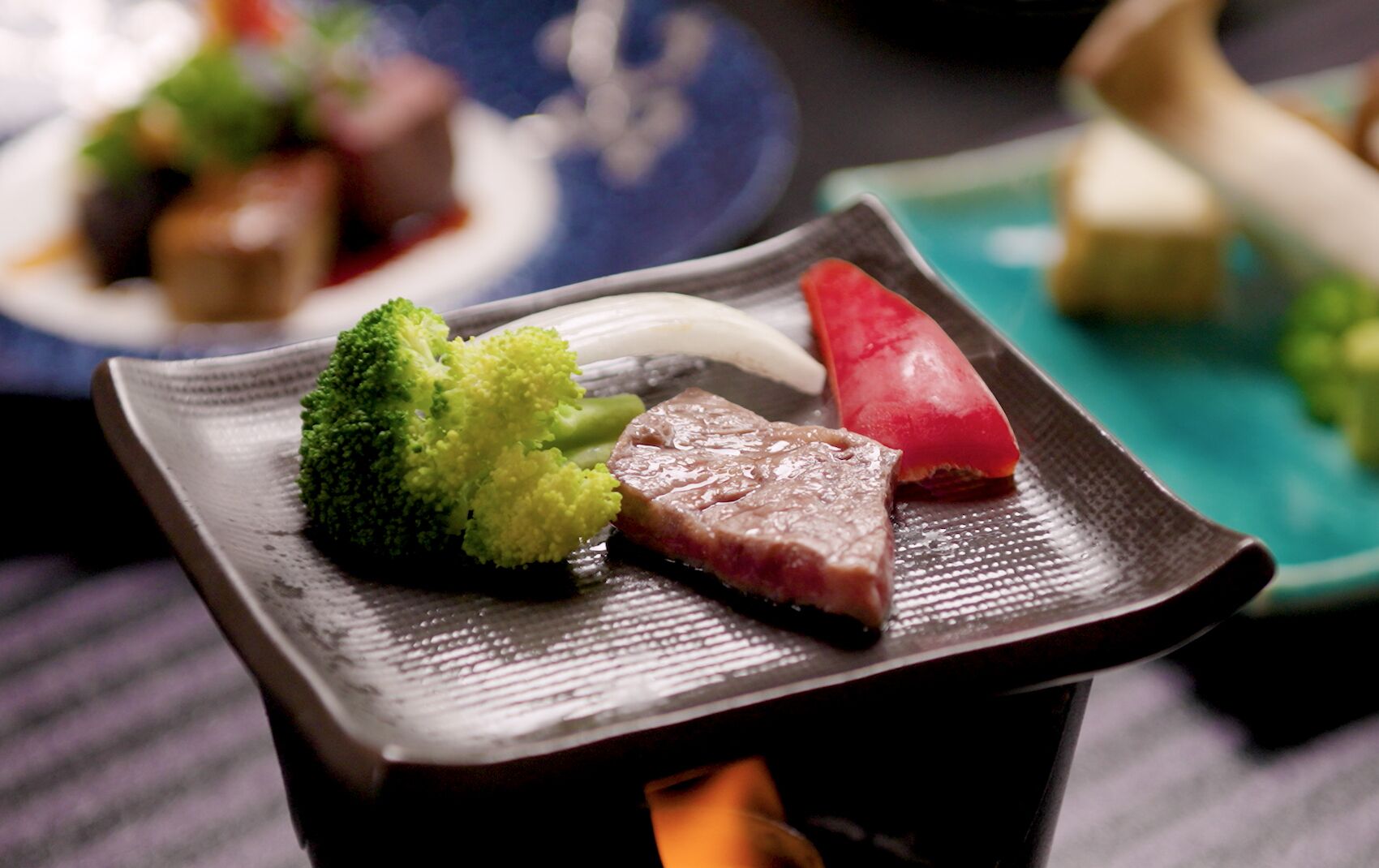 A plate with a piece of meat, a slice of red vegetable, and a broccoli floret on a dark ceramic plate.