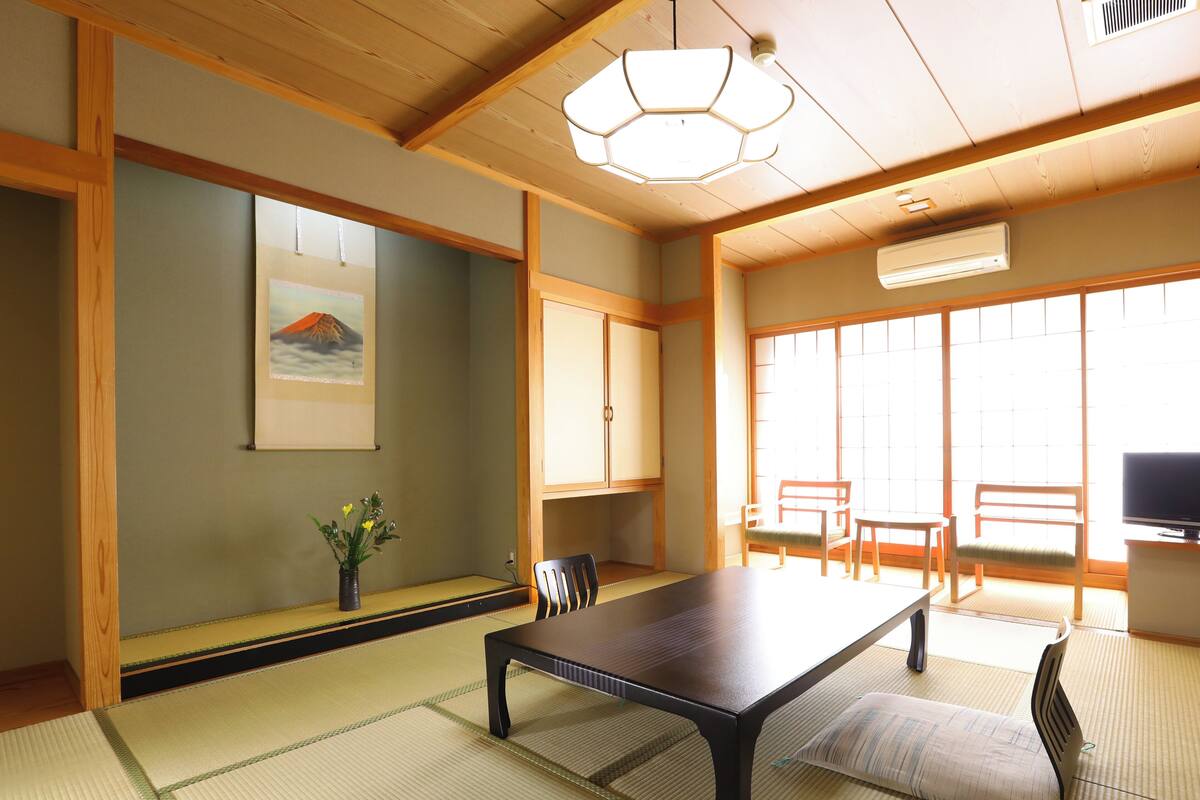 Una habitación japonesa tradicional con suelo de tatami, una mesa baja, sillas de madera, un televisor y un ventanal grande.