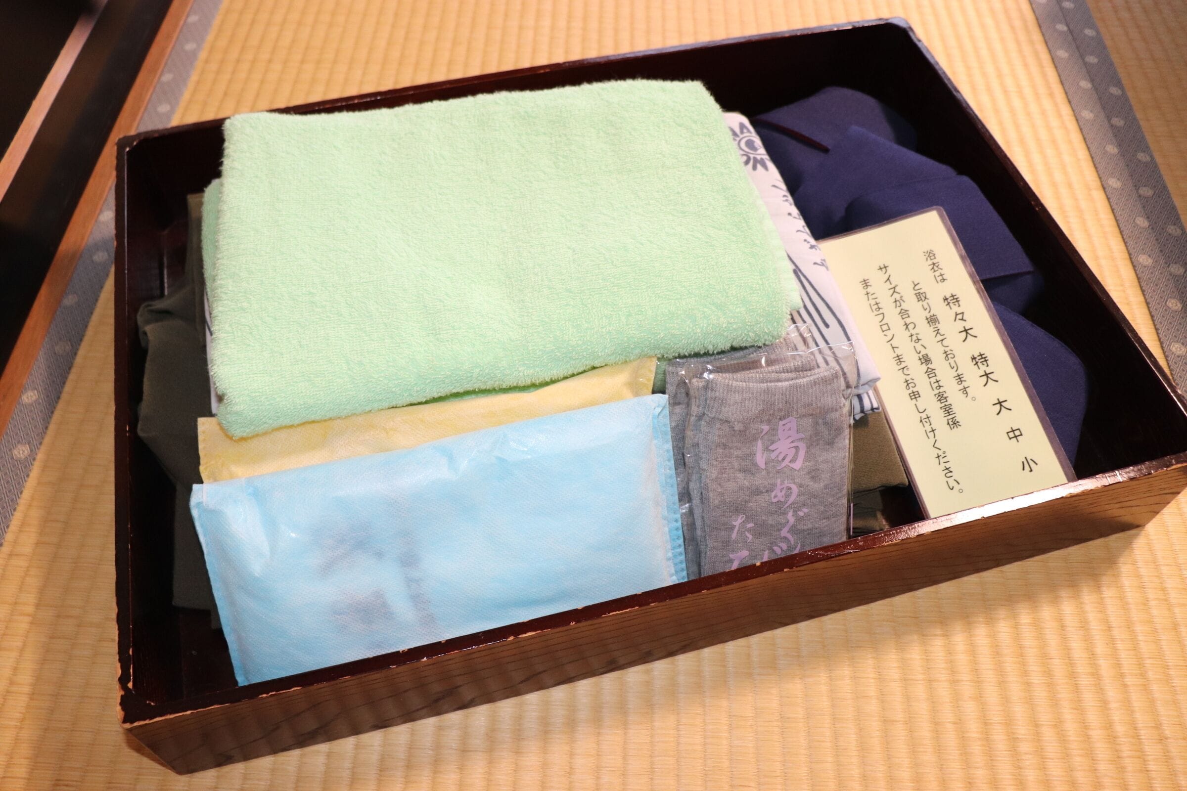 Una caja de madera con varios objetos, incluyendo una toalla verde, una toalla azul y una tarjeta con texto en japonés.